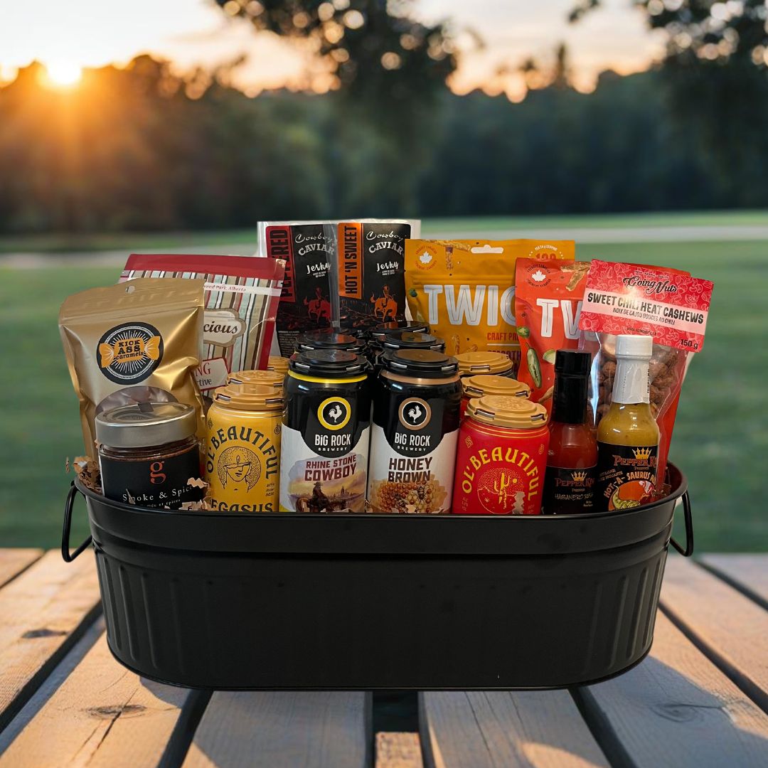 Beer basket featuring local beer cans, jalapeno pretzels, 2 varieties of hot sauce, caramels, chocolate covered coke bottle gummies, gourmet nut mixes and beef jerky. Products sitting in a black metal tin in a park setting. 